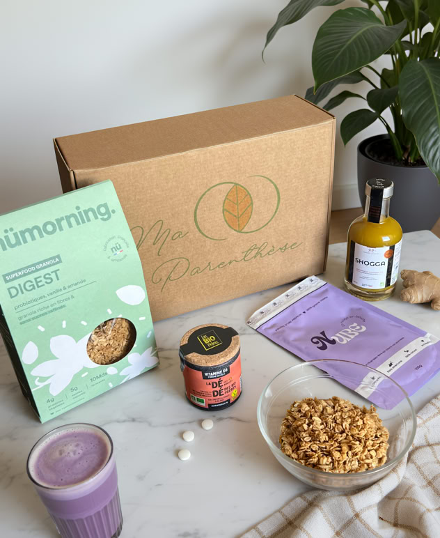 Boîte en carton, paquet de céréales, pot de yaourt, bouteille de jus, verre de smoothie violet, assiette de granola, plante verte en arrière-plan.